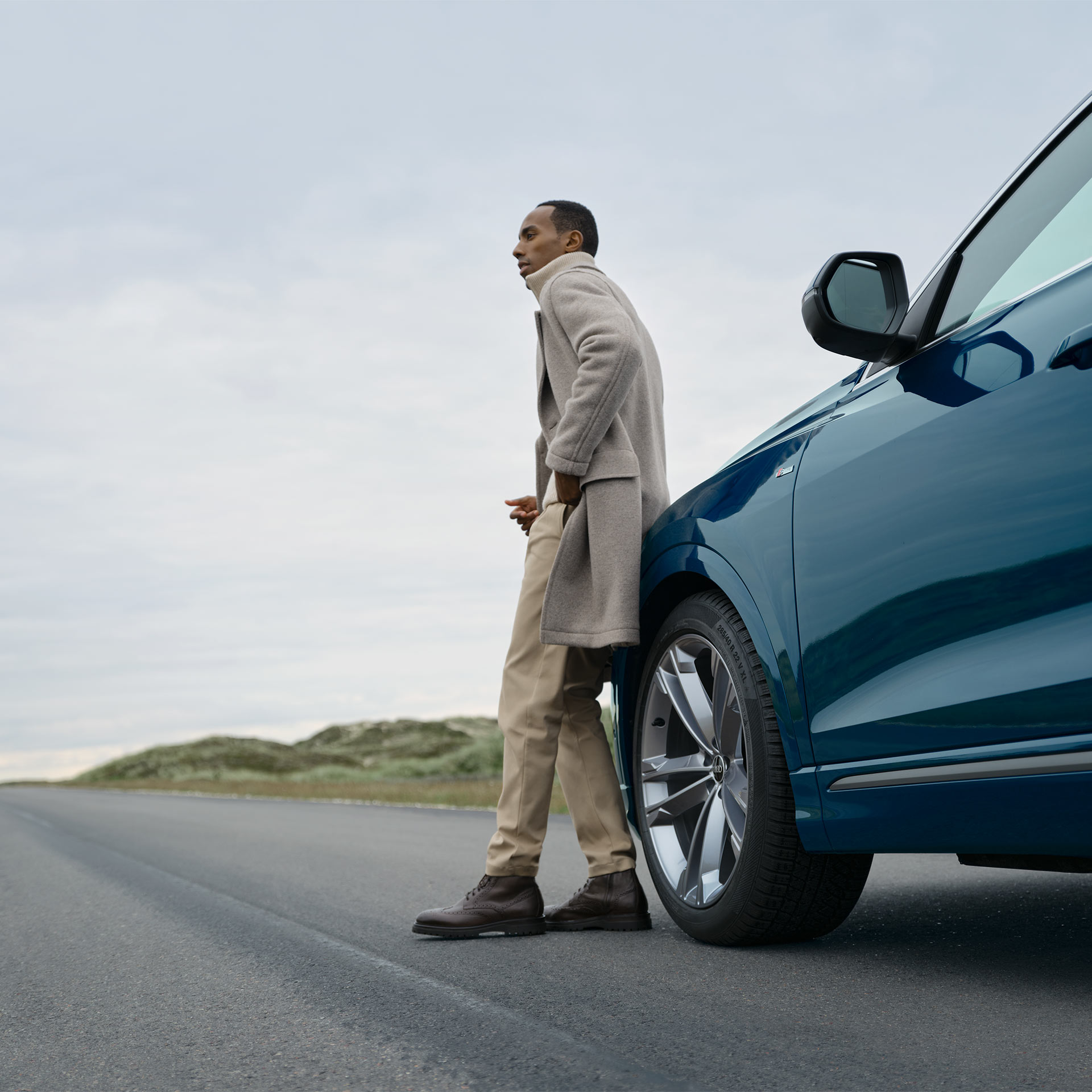A man leaning against the side of a blue Audi parked in the countryside
